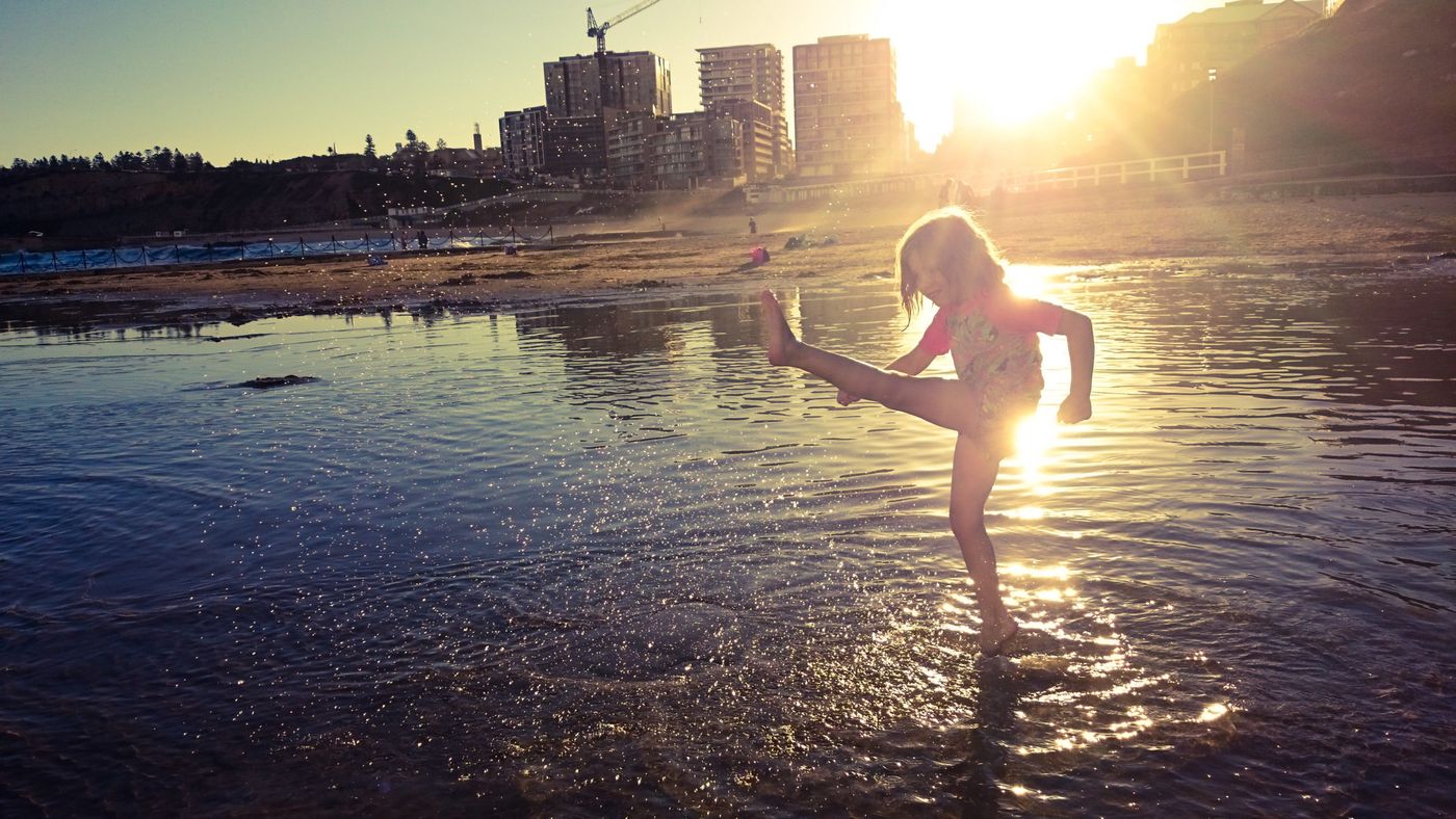 A child kicking up water at Newcastle beach at sunset, with the city skyline behind